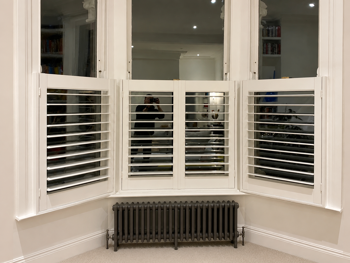Café-style plantation shutters open across a bay window above a period column radiator at night