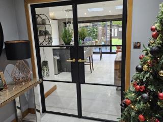 Black steel double doors with brass handles opening from a dining area into a bright kitchen